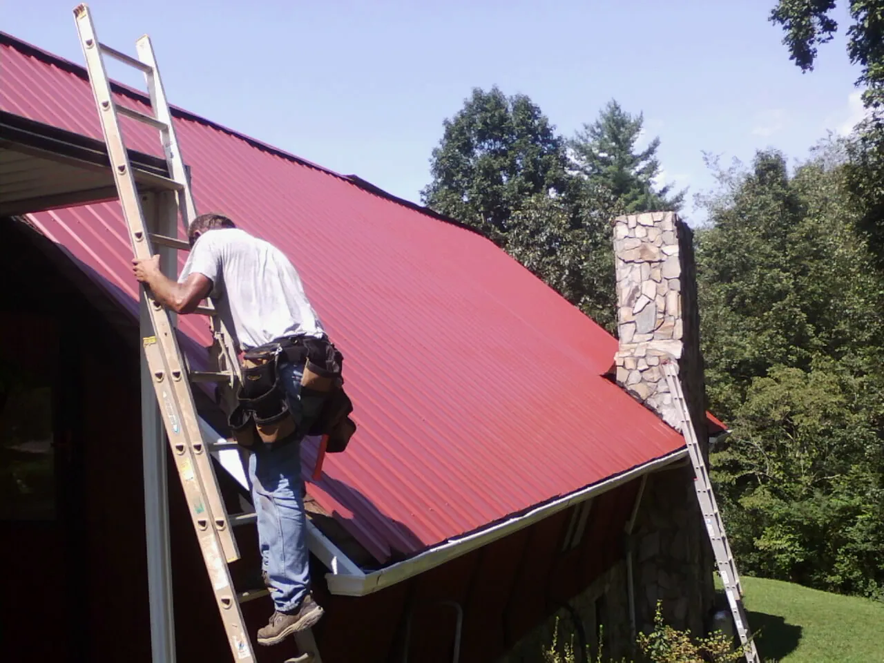 Roofer on a ladder beside a red metal roof and stone chimney — Soffit & Fascia Repair by Roofing Brothers in Tahuya
